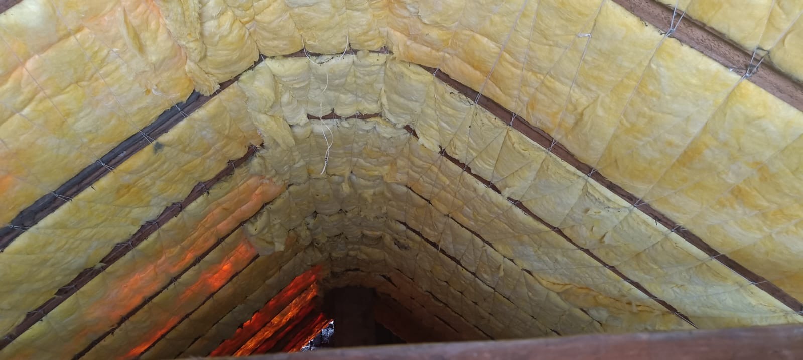 Ocieplenie poddasza wełną mineralną, widok od wewnątrz. Żółta wełna między krokwiami, zabezpieczona sznurkiem. Ujęcie z dołu, w kierunku szczytu dachu.