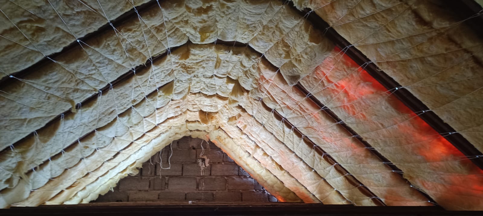 Ocieplenie poddasza wełną mineralną, widok od dołu. Widoczne krokwie i siatka podtrzymująca izolację. Fragment ściany z cegieł. Czerwone światło w tle.