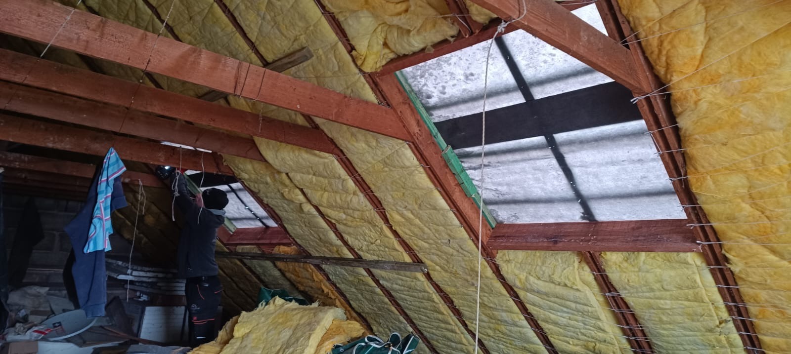 Ocieplenie poddasza wełną mineralną, widoczne drewniane krokwie i okno dachowe. Prace izolacyjne w toku, widoczny pracownik w trakcie montażu.