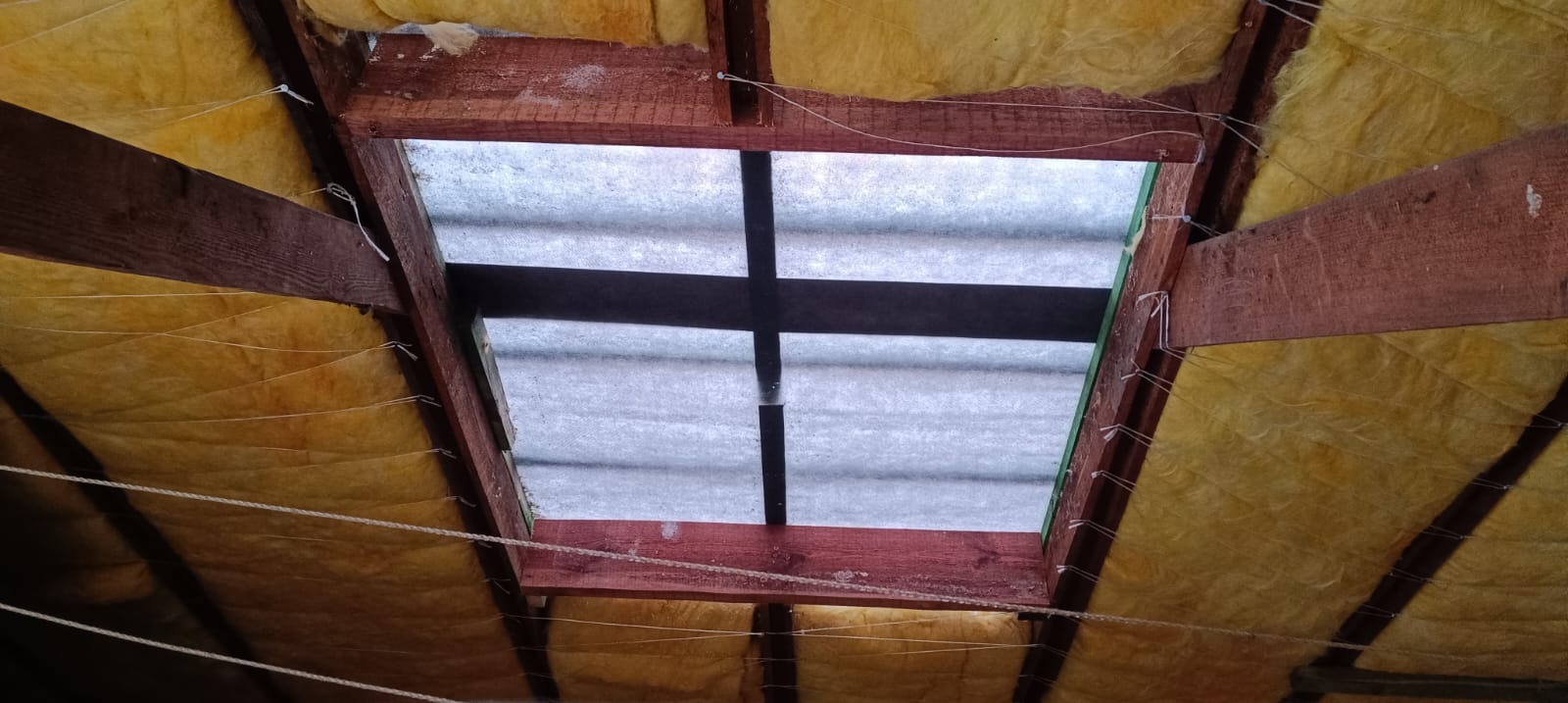 Ocieplenie poddasza wełną mineralną, widok od wewnątrz. Drewniana konstrukcja dachu z izolacją, zabezpieczona sznurkiem. Fragment okna dachowego.