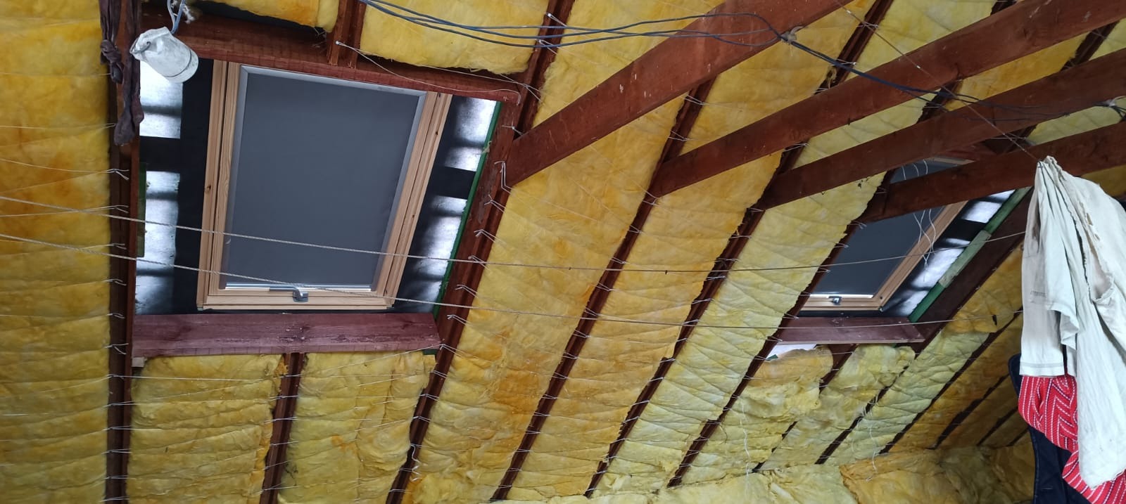 Ocieplenie poddasza wełną mineralną, widoczne drewniane krokwie i okna dachowe. Prace wykończeniowe w trakcie realizacji, widoczne przewody i ubrania robocze.