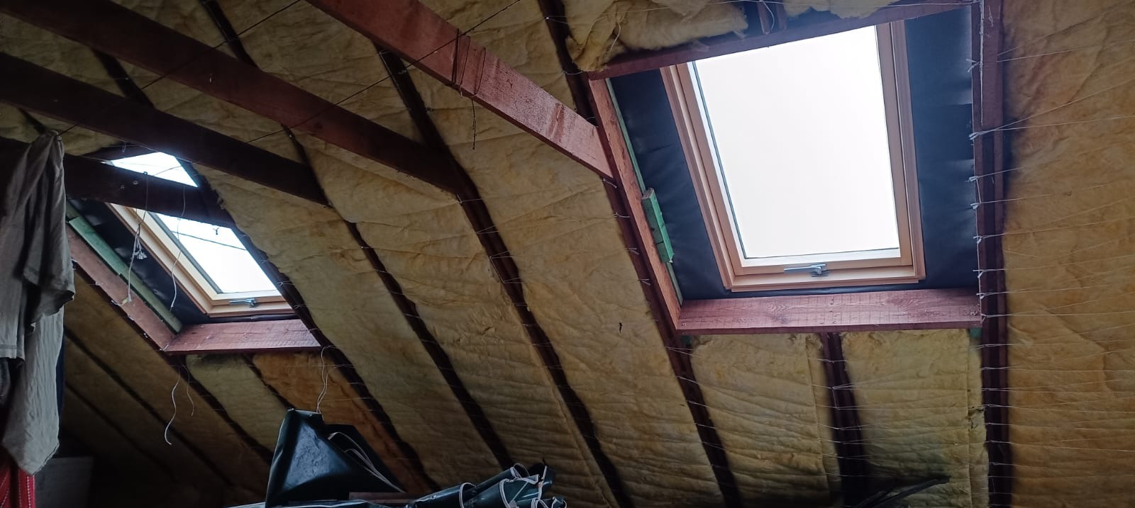 Ocieplenie poddasza wełną mineralną, widoczne drewniane krokwie i dwa okna dachowe. Prace wykończeniowe w trakcie realizacji.