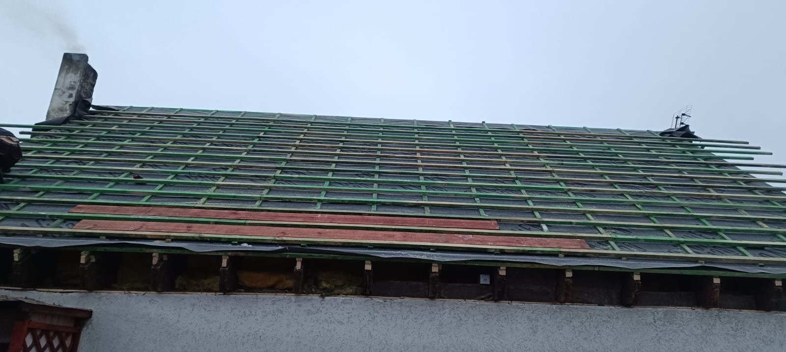 Konstrukcja dachu z łatami i membraną, widoczny komin i antena. Częściowo odsłonięta izolacja. Etap prac przed położeniem pokrycia dachowego.