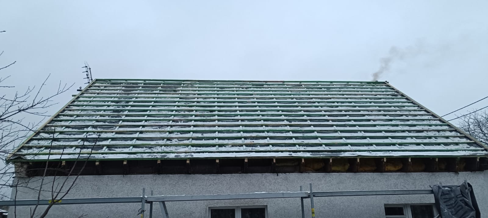 Dach w trakcie prac: widoczne łaty i kontrłaty, częściowo pokryte śniegiem. Fragment ściany budynku z oknem i rusztowaniem. W tle drzewa i pochmurne niebo.