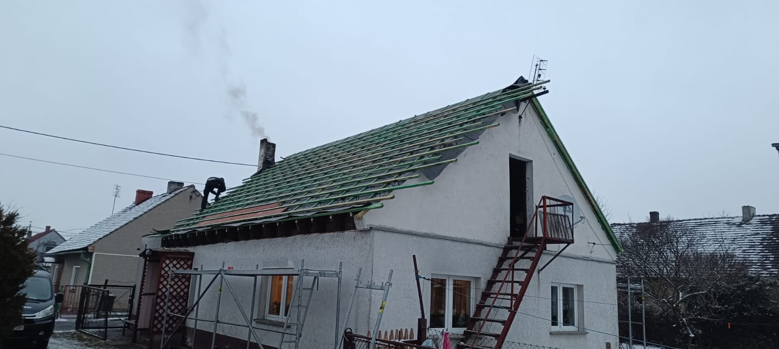 Prace na dachu domu jednorodzinnego: dekarz montuje konstrukcję pod nowe pokrycie, widoczne rusztowania i komin z dymem. Ujęcie z dołu, pochmurny dzień.