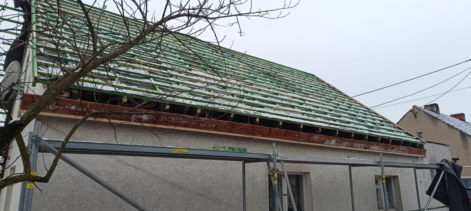 Konstrukcja dachu z łatami i membraną, widoczna z rusztowania. Fragment budynku z oknami i drzewo z przodu. Zimowa sceneria z lekkim śniegiem na dachu.