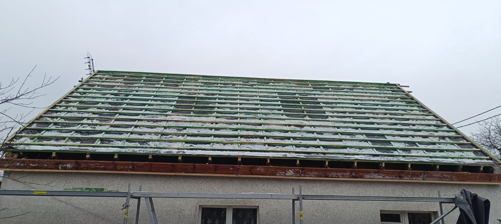 Dach w trakcie prac: widoczne łaty, membrana i fragment ośnieżonej konstrukcji. Na pierwszym planie fragment ściany budynku i rusztowanie.