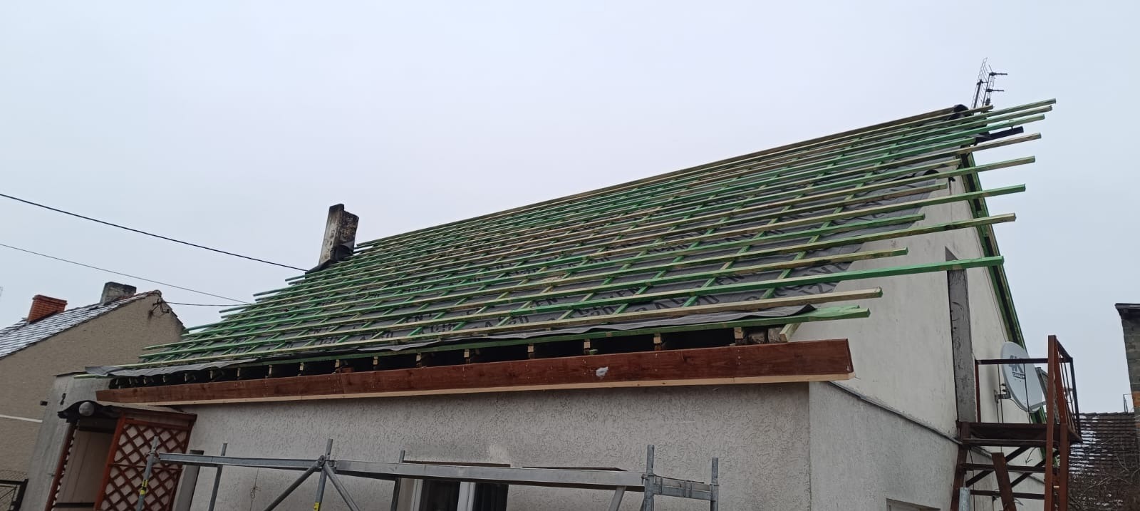 Konstrukcja dachu z łatami i membraną, widok z dołu. Elementy drewniane w kolorze zielonym. Prace dekarskie w toku, widoczny komin i antena satelitarna.