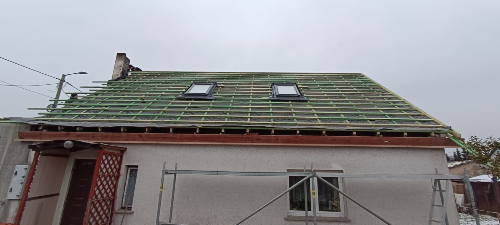 Montaż łat na dachu domu jednorodzinnego z dwoma oknami dachowymi. Widoczne rusztowanie i pracownik na dachu. Etap przygotowania pod pokrycie.