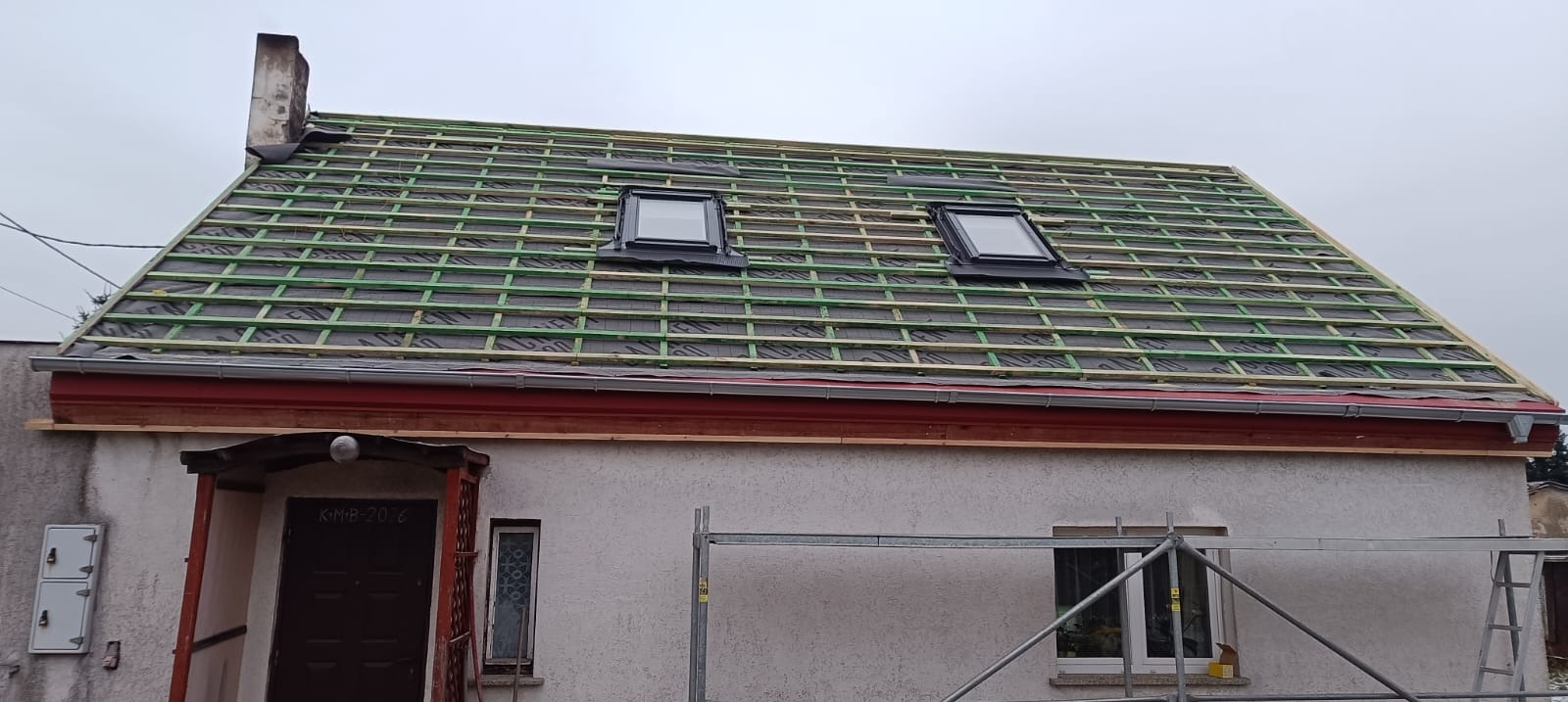 Dach w trakcie wymiany: widoczne łaty, membrana i okna dachowe. Ujęcie z dołu, na pierwszym planie fragment elewacji budynku i rusztowanie.