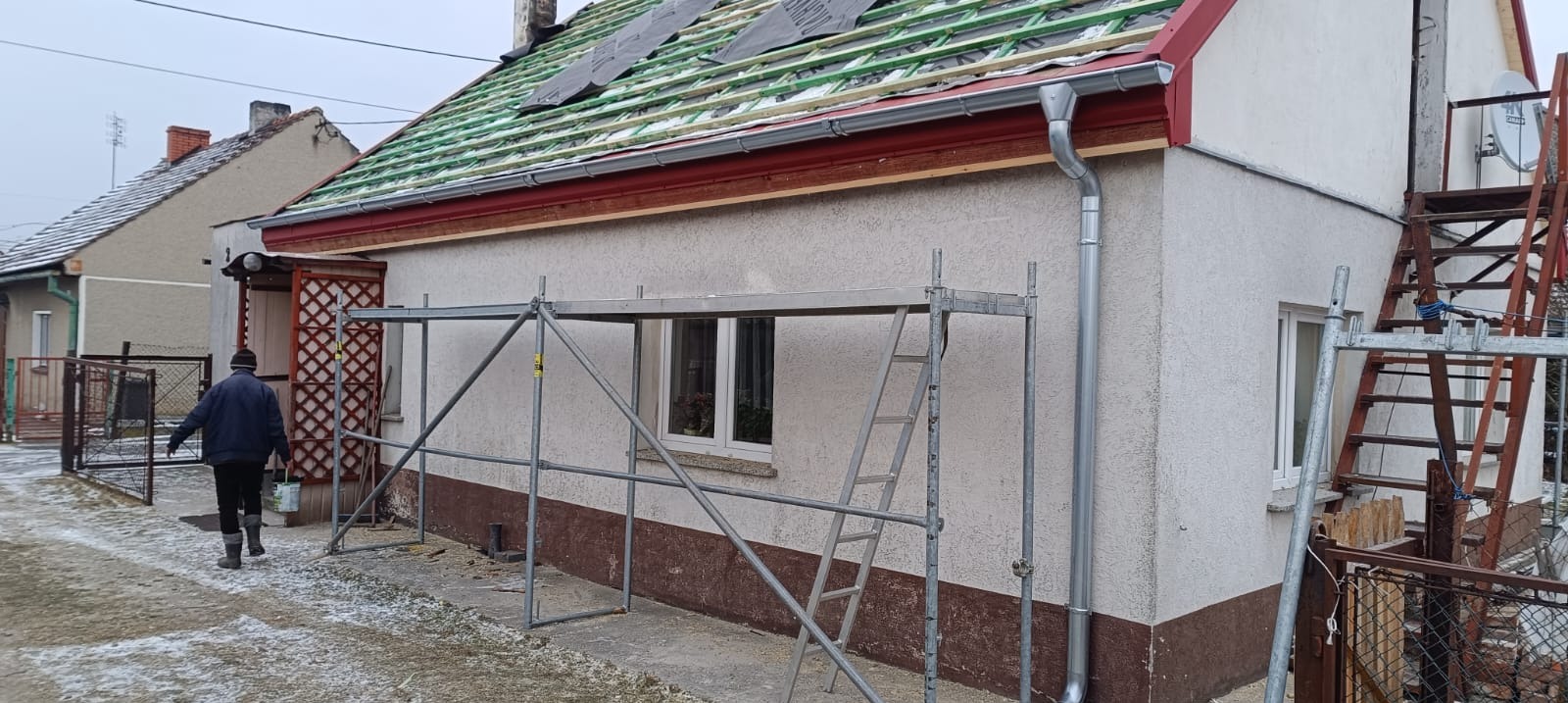 Dom jednorodzinny w trakcie prac dekarskich: widoczne rusztowania, nowa podkonstrukcja dachu z łatami i fragmenty papy. Zamontowane rynny. Prace w toku.