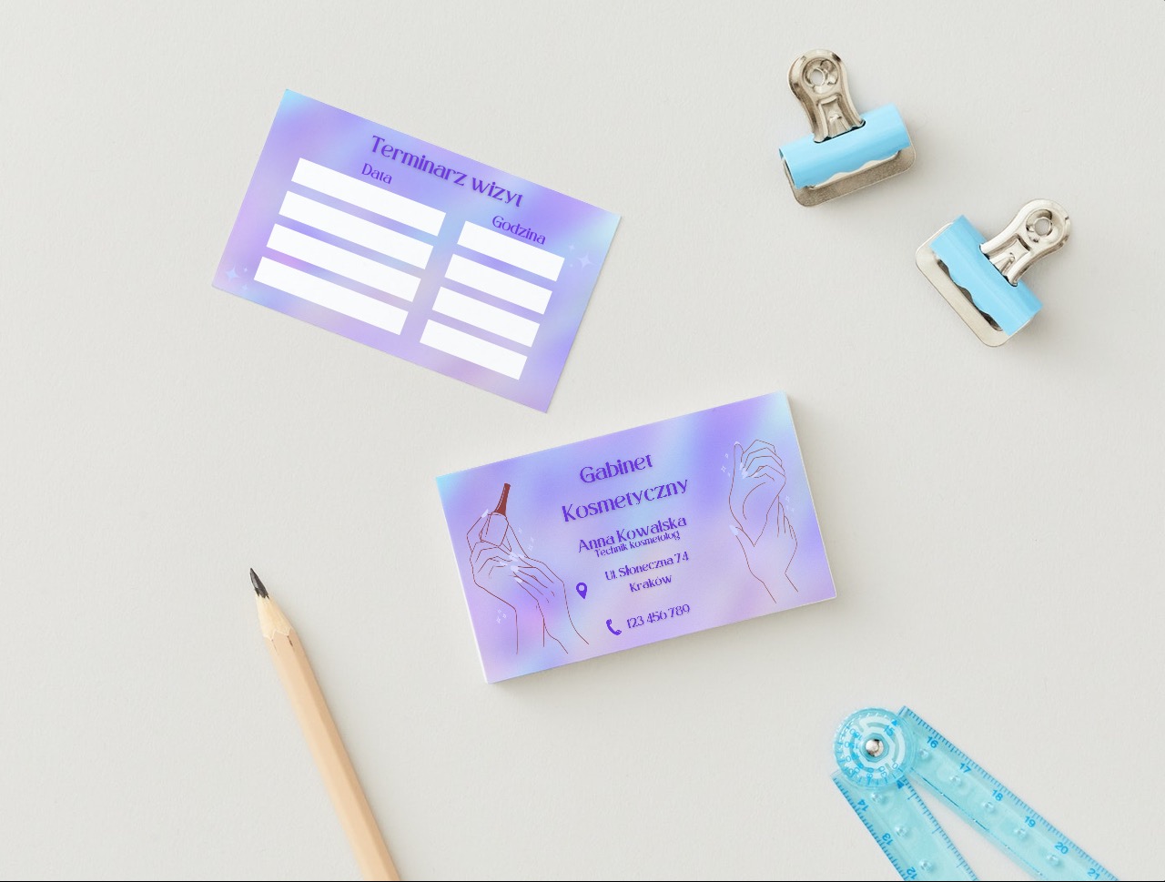 Dwie wizytówki w pastelowych kolorach z elementami graficznymi, leżące na białym tle obok ołówka, niebieskiej linijki i dwóch spinaczy biurowych.