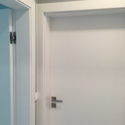 An-Door Serwis Autoryzowany Montaż Drzwi Systemu PORTA - Dwa białe skrzydła drzwiowe z ościeżnicami, widoczne zawiasy ukryte w jednym skrzydle, klamka z prostokątnym szyldem w drugim skrzydle.