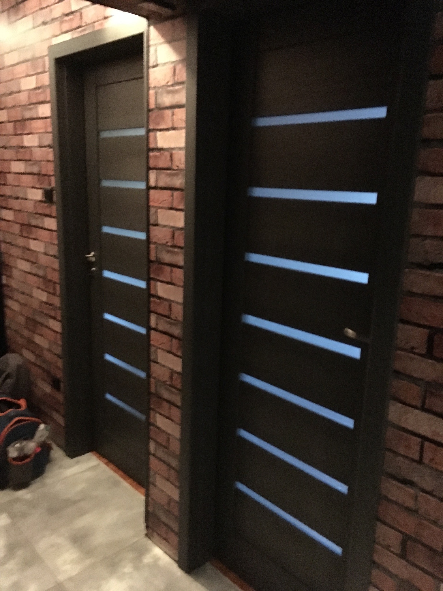 Dwie nowoczesne, ciemne drzwi wewnętrzne z poziomymi przeszkleniami, zamontowane w ścianie z cegły, widok z boku.