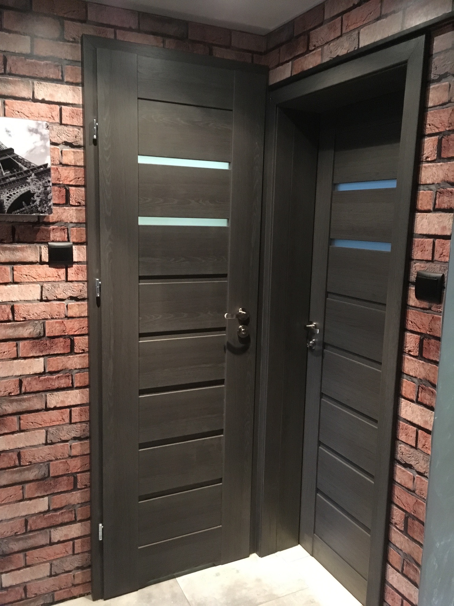 Dwie nowoczesne, ciemne, drewniane drzwi z poziomymi przeszkleniami, zamontowane w rogu pokoju z ceglaną ścianą i wiszącym obrazem wieży Eiffla.