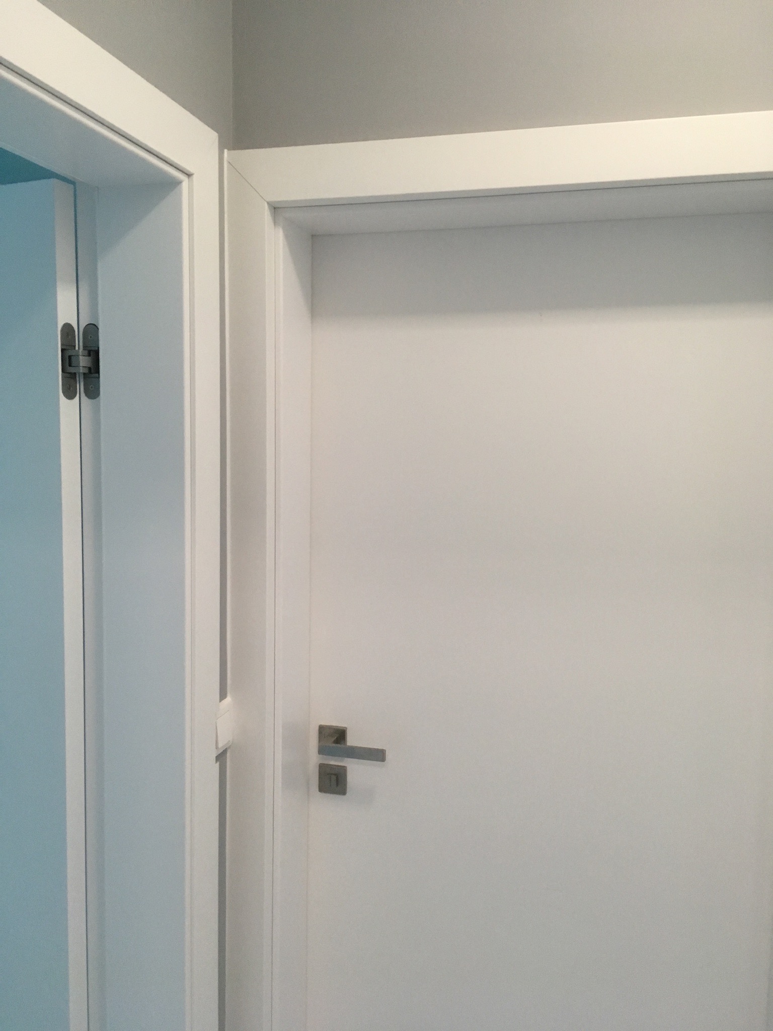 Dwa białe skrzydła drzwiowe z ościeżnicami, widoczne zawiasy ukryte w jednym skrzydle, klamka z prostokątnym szyldem w drugim skrzydle.