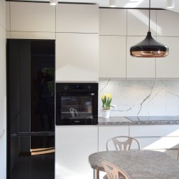 MODERN STUDIO W.FIRLEJ, M.FIRLEJ Spółka cywilna - Nowoczesna kuchnia z białymi szafkami, czarnym piekarnikiem i lodówką, marmurowym blatem, lampą wiszącą w kolorze miedzi i okrągłym stołem z krzesłami z kwiatowym wzorem na siedziskach.