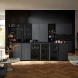 Nowoczesna kuchnia z ciemnymi, drewnianymi szafkami, szklanymi witrynami i czarną ścianą, uzupełniona oświetleniem i eleganckim salonem z brązową sofą.