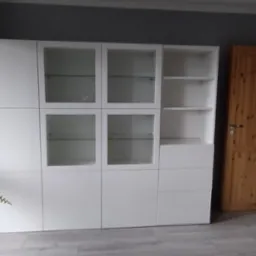 Biała, modułowa szafa z przeszklonymi drzwiczkami, stojąca na panelach podłogowych w pokoju z szarą ścianą i drewnianymi drzwiami, obok okna z widoczną zielenią.