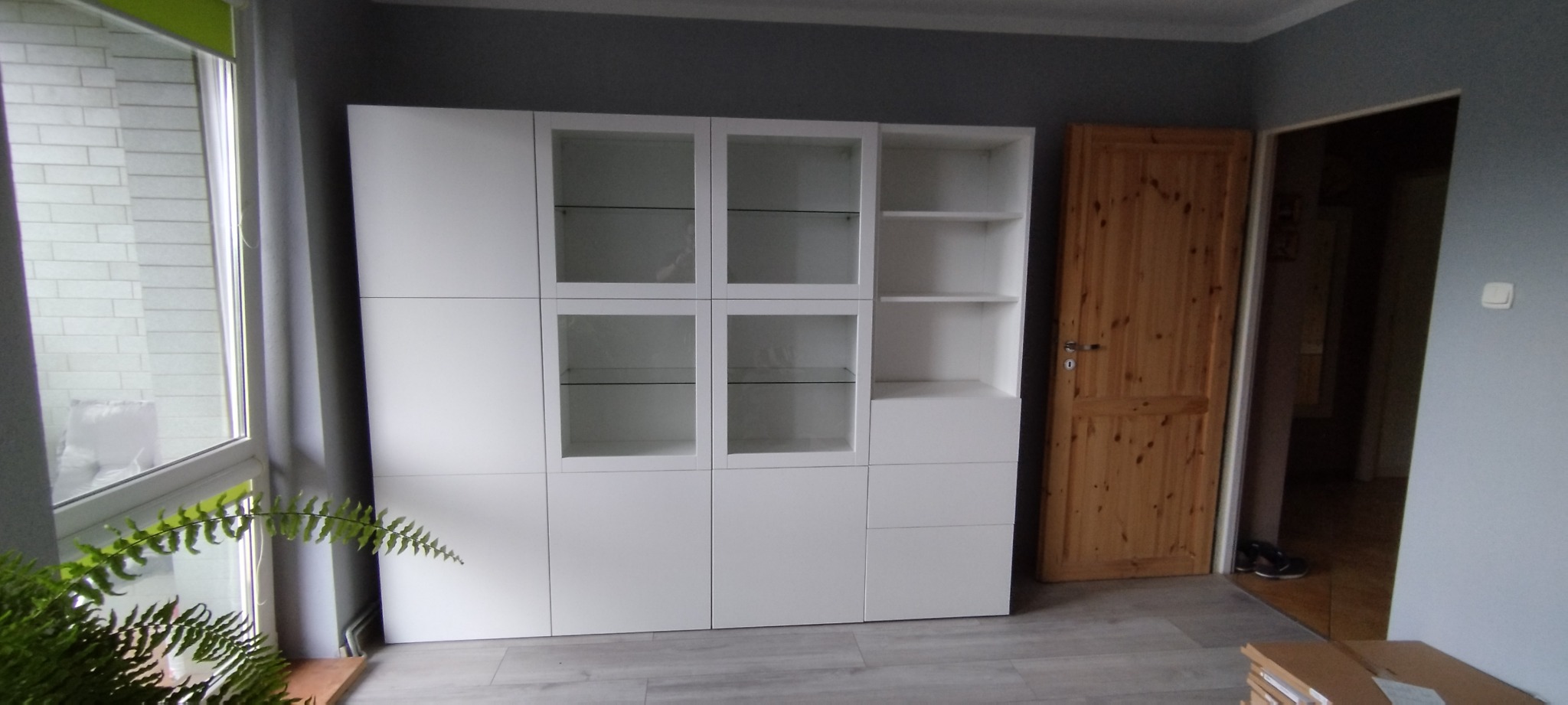 Biała, modułowa szafa z przeszklonymi drzwiczkami, stojąca na panelach podłogowych w pokoju z szarą ścianą i drewnianymi drzwiami, obok okna z widoczną zielenią.