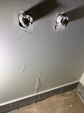 Szara ściana z dwoma otworami na gniazdka elektryczne, wypełnionymi białym materiałem izolacyjnym, widoczne ślady po niedokładnym szpachlowaniu, szara listwa przypodłogowa i zanieczyszczona podłoga.