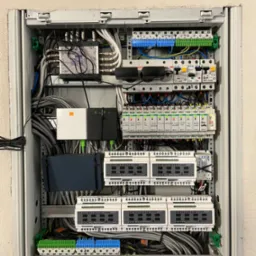 Otwarta szafka rozdzielcza z licznymi przewodami, modułami sterującymi i zabezpieczeniami, w tym moduły Satel, router Orange i rozgałęźnik sygnału satelitarnego.