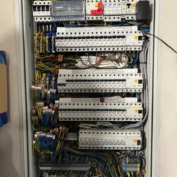 Rozdzielnica elektryczna z licznymi bezpiecznikami, przewodami i elementami sterującymi, widoczne oznaczenia na przewodach i komponentach, marki Legrand i Eaton.