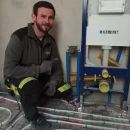 Instalator w szarym polarze PSB, wskazujący palcem na stelaż podtynkowy Geberit z niebieskimi rurami przy ogrzewaniu podłogowym z białych rurek na czerwonej macie, w surowym wnętrzu.