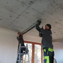 Dwóch instalatorów na drabinach montuje rury wentylacyjne pod betonowym sufitem w Wielowsi, jeden z nich pokazuje kciuk w górę.