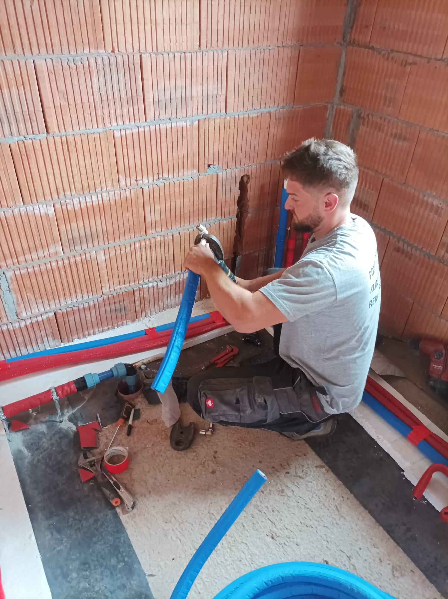 Instalator w szarej koszulce łączy niebieską rurę instalacyjną w surowym wnętrzu z ceglanymi ścianami, używając zaciskarki; widoczne narzędzia, czerwone i niebieskie rury oraz wiercąca wiertarka...