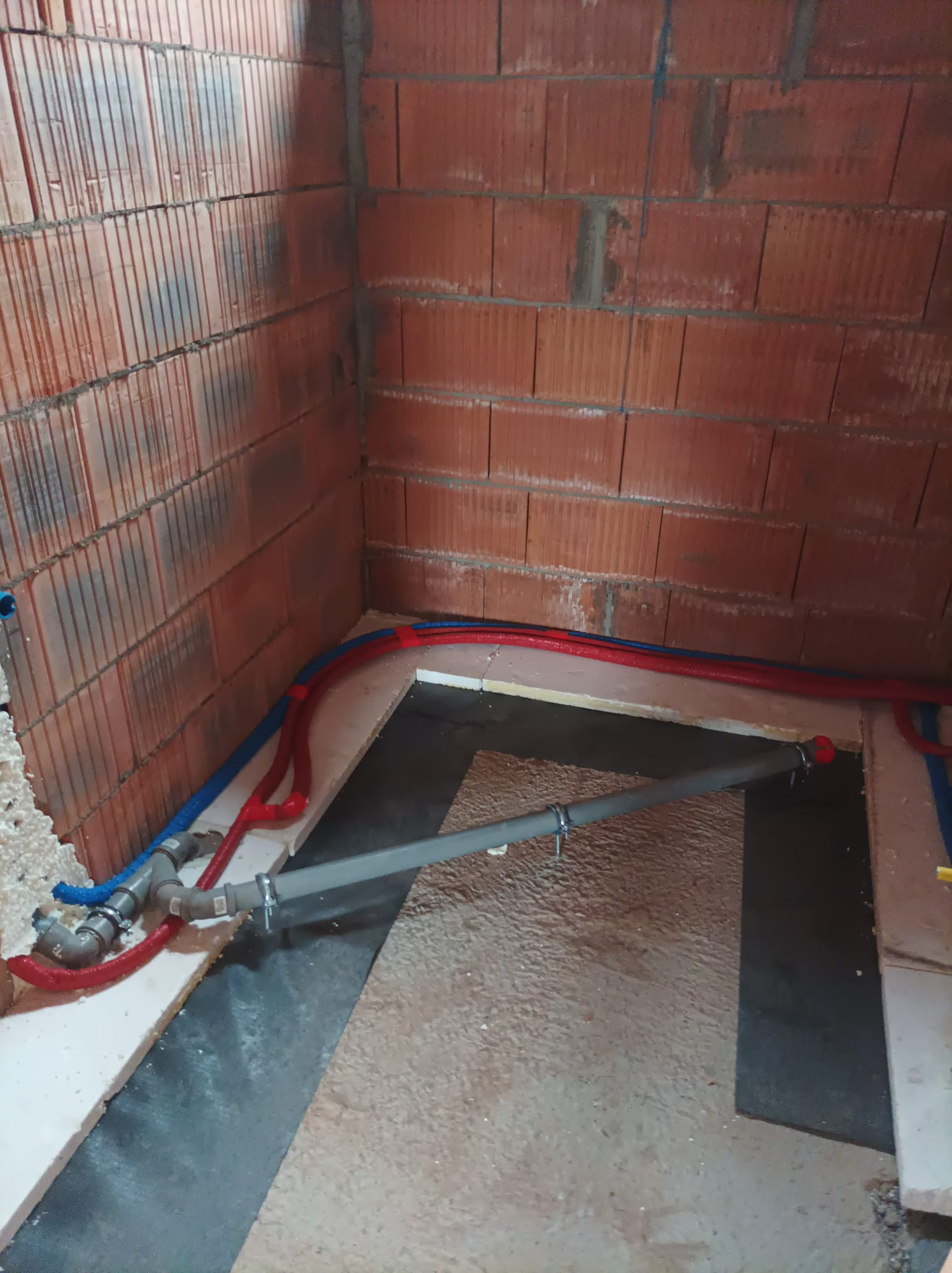 Instalacja ogrzewania podłogowego w trakcie montażu: widoczne rury w kolorze czerwonym i niebieskim, mocowane do podłoża, z surowymi ścianami z czerwonej cegły w tle.