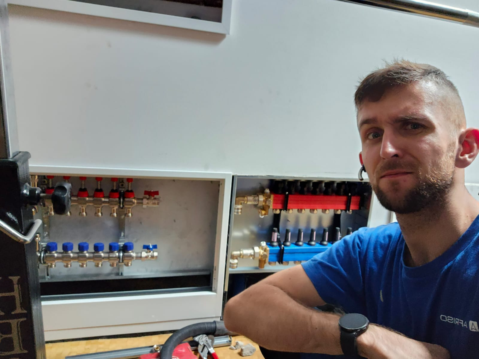 Mężczyzna w niebieskiej koszulce prezentuje rozdzielacz ogrzewania podłogowego z czerwonymi i niebieskimi zaworami, zamontowany w białej szafce.