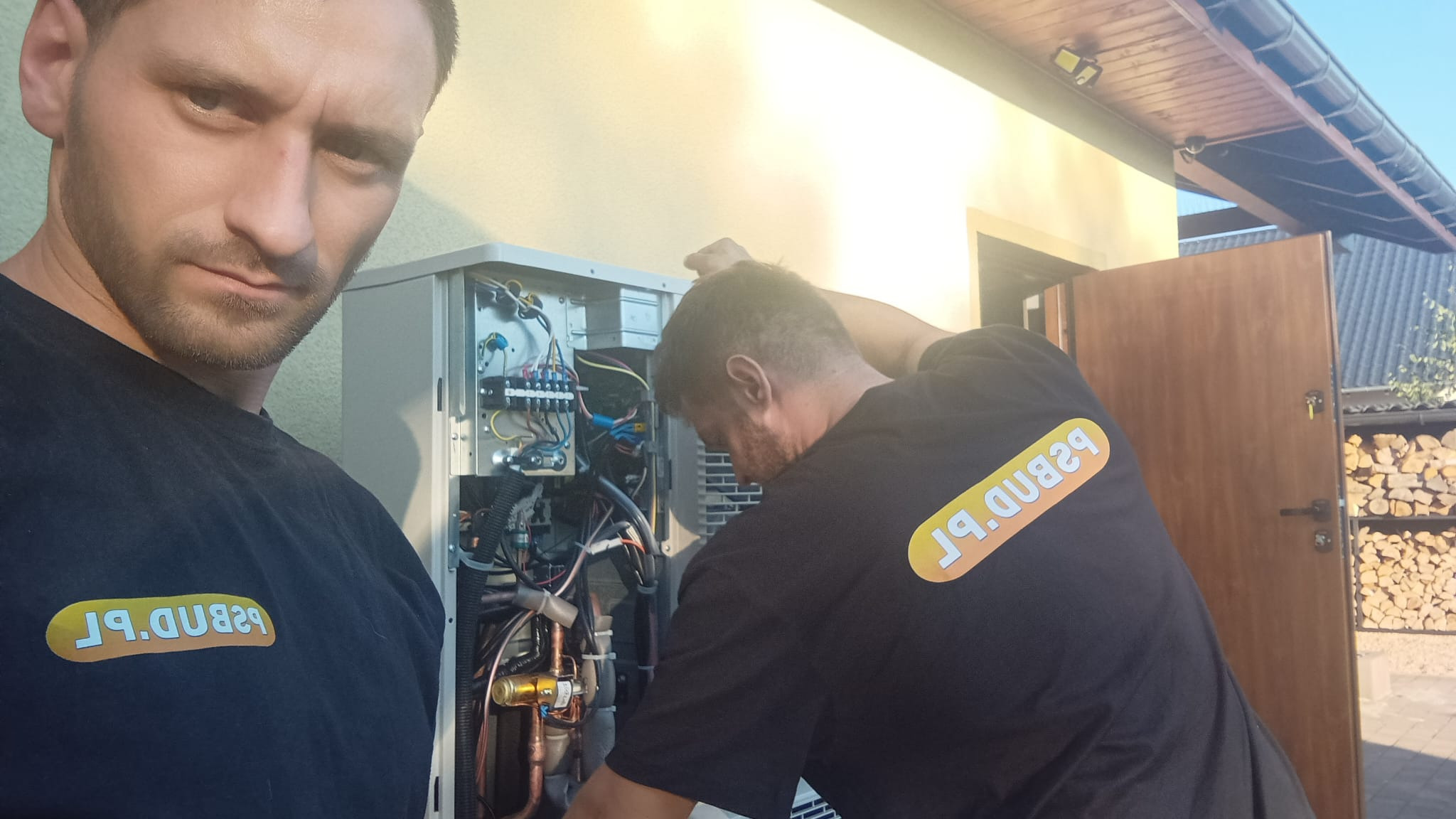 Dwóch instalatorów w czarnych koszulkach z logo firmy, jeden z przodu z poważnym wyrazem twarzy, drugi w trakcie podłączania przewodów wewnątrz otwartej obudowy urządzenia grzewczego na zewnątrz...