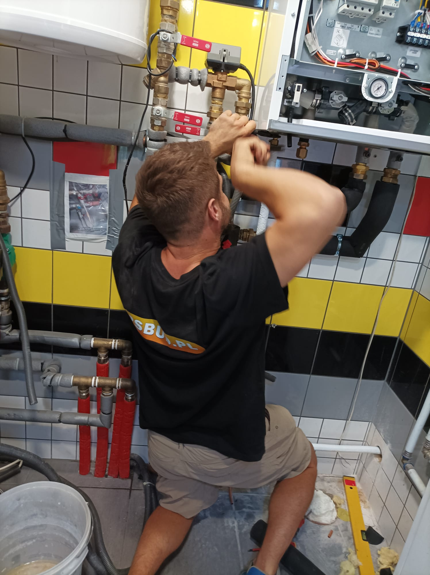 Instalator w trakcie montażu kotła grzewczego w pomieszczeniu z biało-żółto-czarnymi płytkami, widoczne rury, zawory i elementy elektryczne, na koszulce logo firmy.