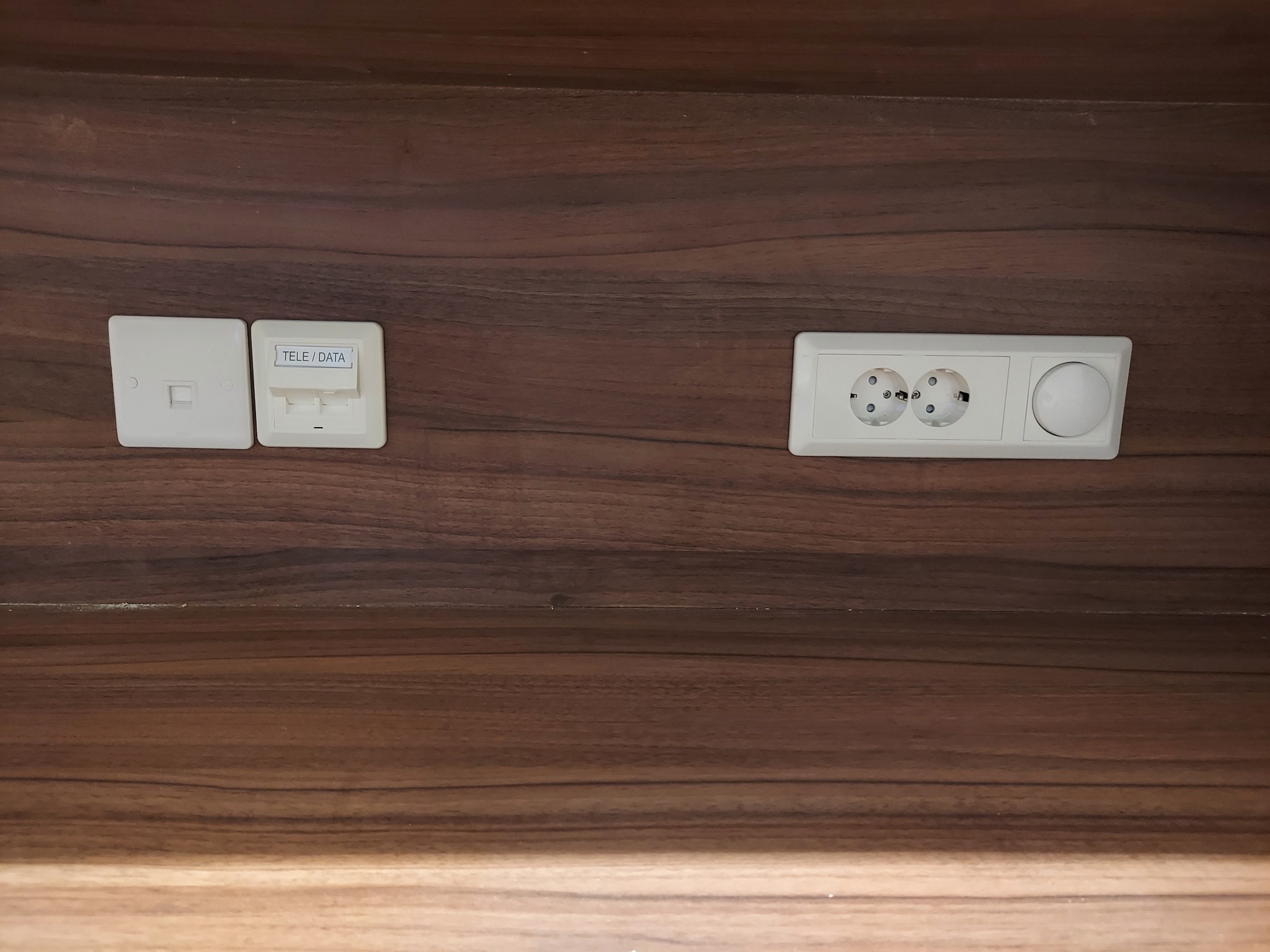Gniazdka elektryczne i gniazdo teleinformatyczne zamontowane w ciemnobrązowej drewnianej ścianie, widoczne z bliska.