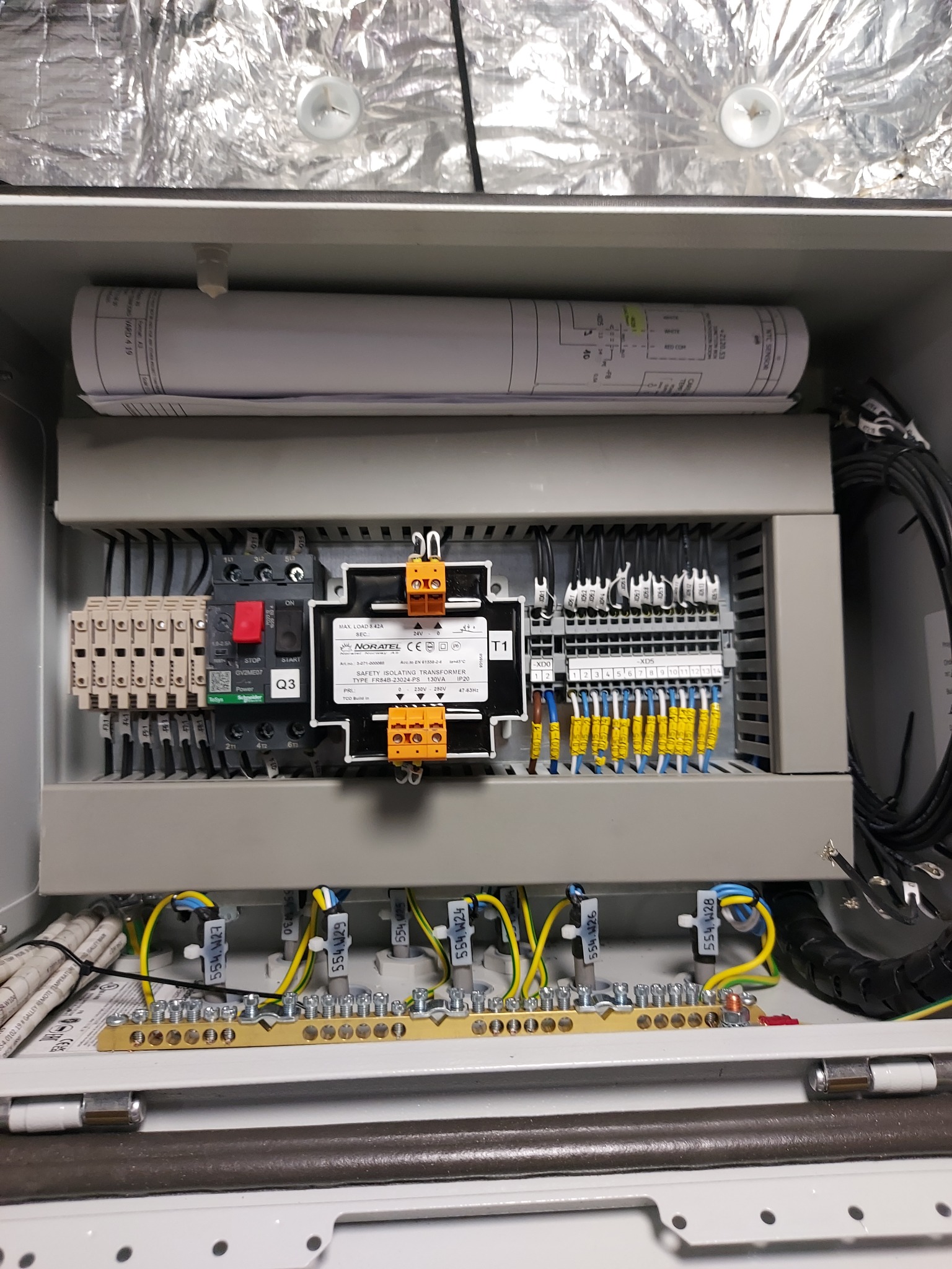 Wnętrze szafy sterowniczej z transformatorem izolacyjnym, bezpiecznikami, listwami zaciskowymi i okablowaniem, widoczne schematy elektryczne na rolce papieru.