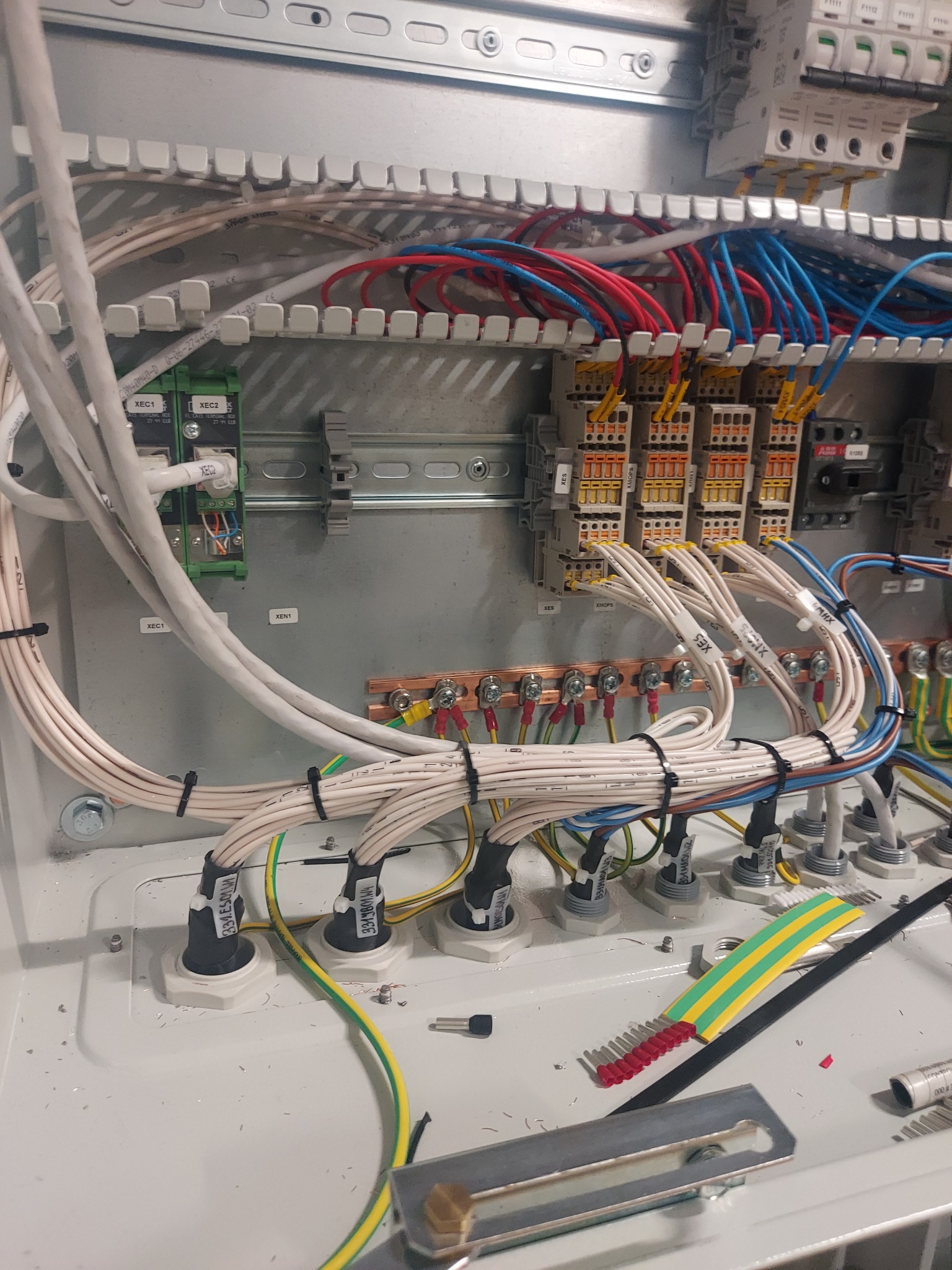 Wnętrze szafy elektrycznej z widocznymi przewodami o różnych kolorach, listwami zaciskowymi, zabezpieczeniami i elementami montażowymi, częściowo okablowane, z narzędziami i elementami...