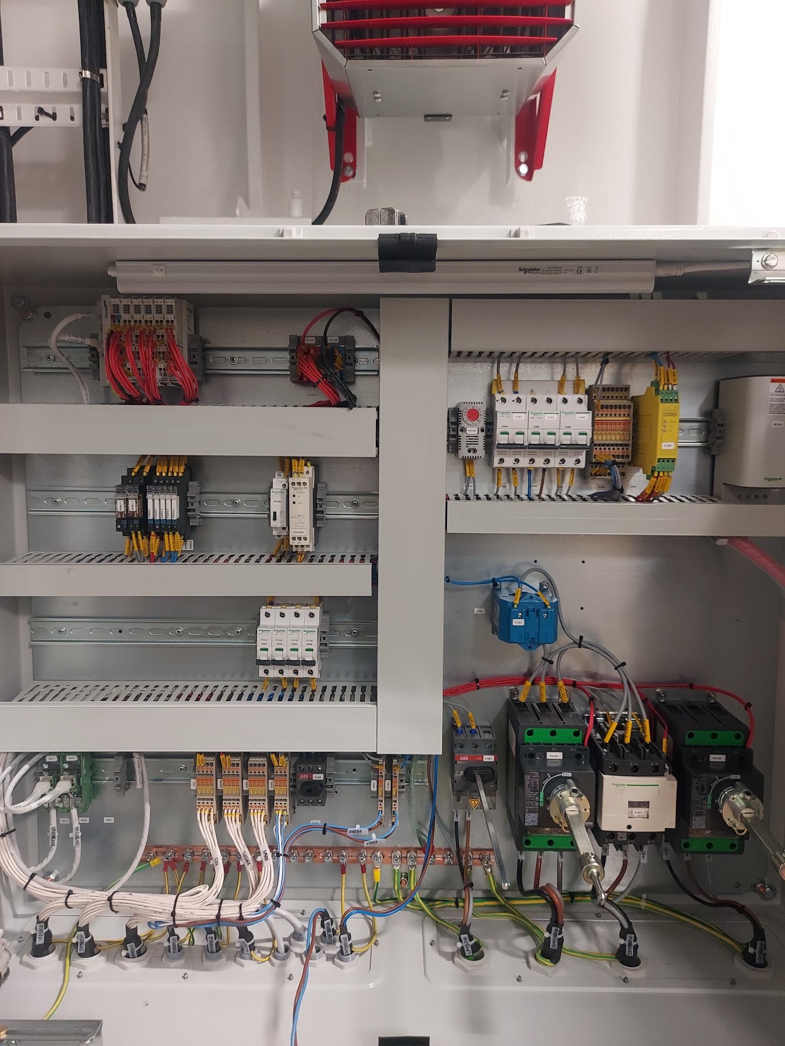 Otwarta szafa elektryczna z widocznymi bezpiecznikami, stycznikami, przewodami oznaczonymi etykietami i szynami DIN, zainstalowana w pomieszczeniu technicznym z czerwonym elementem wentylacyjnym u...