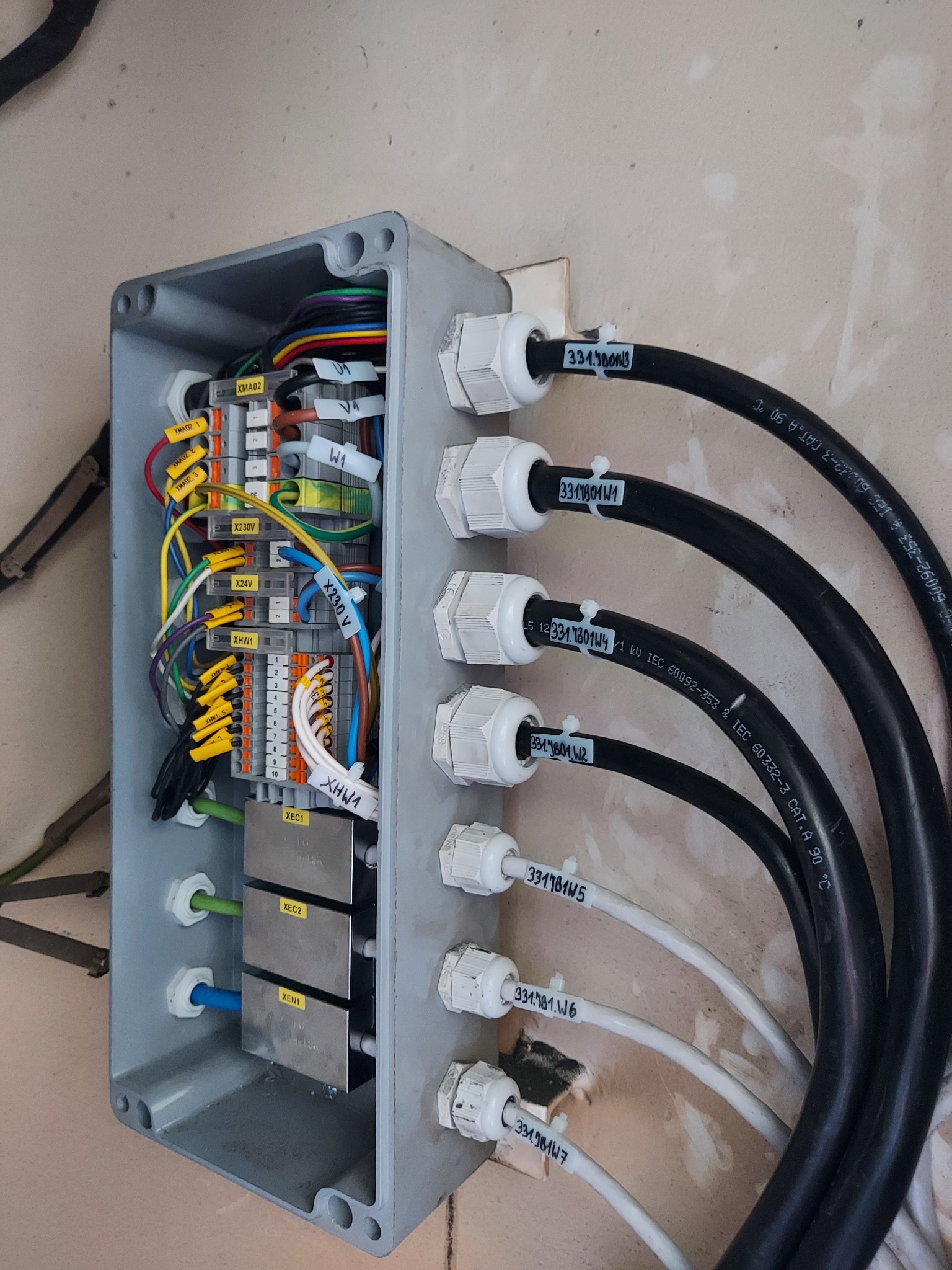 Otwarta szara skrzynka rozdzielcza z widocznymi kablami o różnych kolorach, złączkami i oznaczeniami, zamontowana na ścianie z widocznymi śladami po malowaniu.