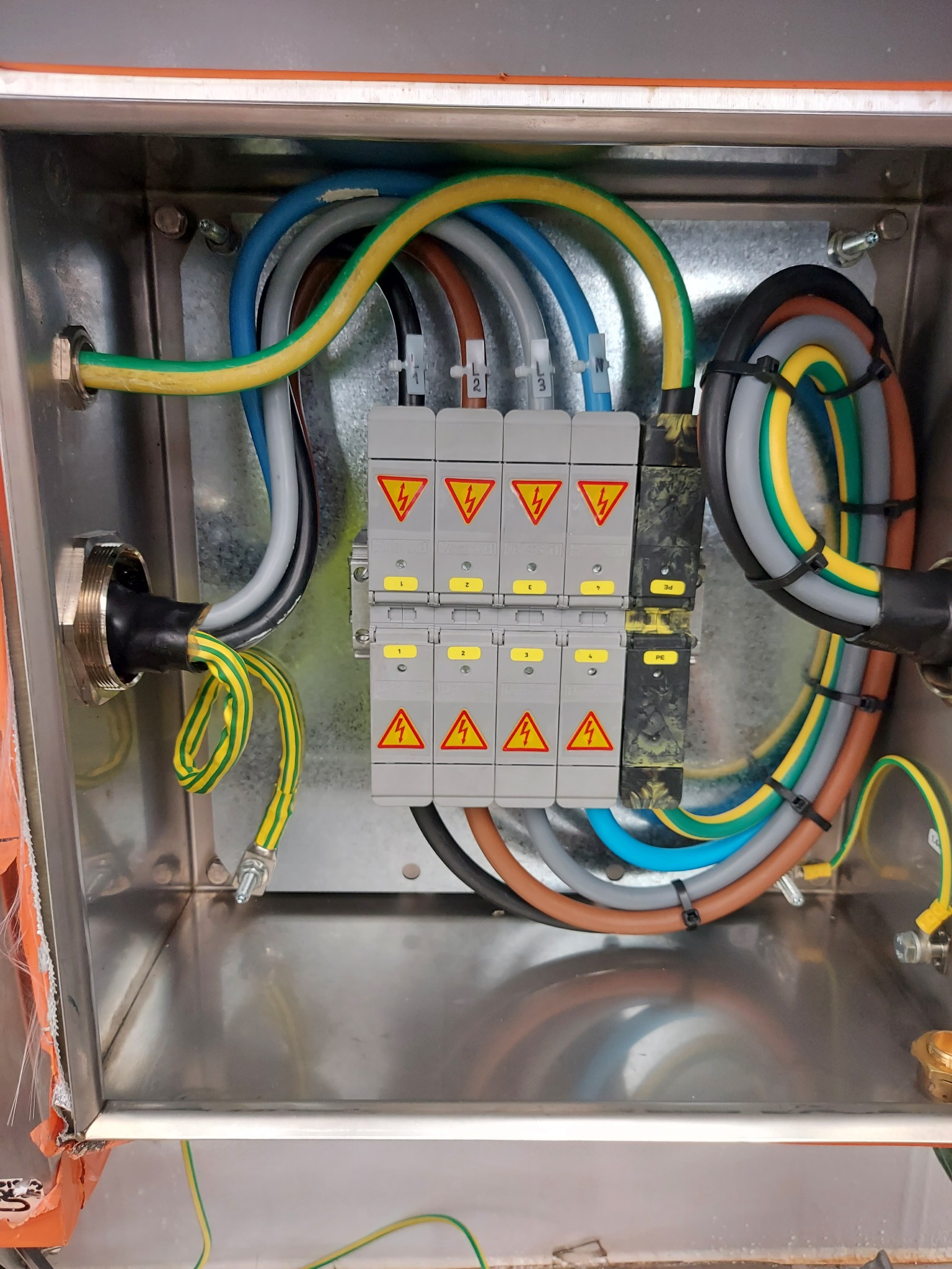 Wnętrze metalowej skrzynki elektrycznej z zamontowanymi bezpiecznikami i uporządkowanymi kablami o różnych kolorach, zabezpieczonymi opaskami zaciskowymi.