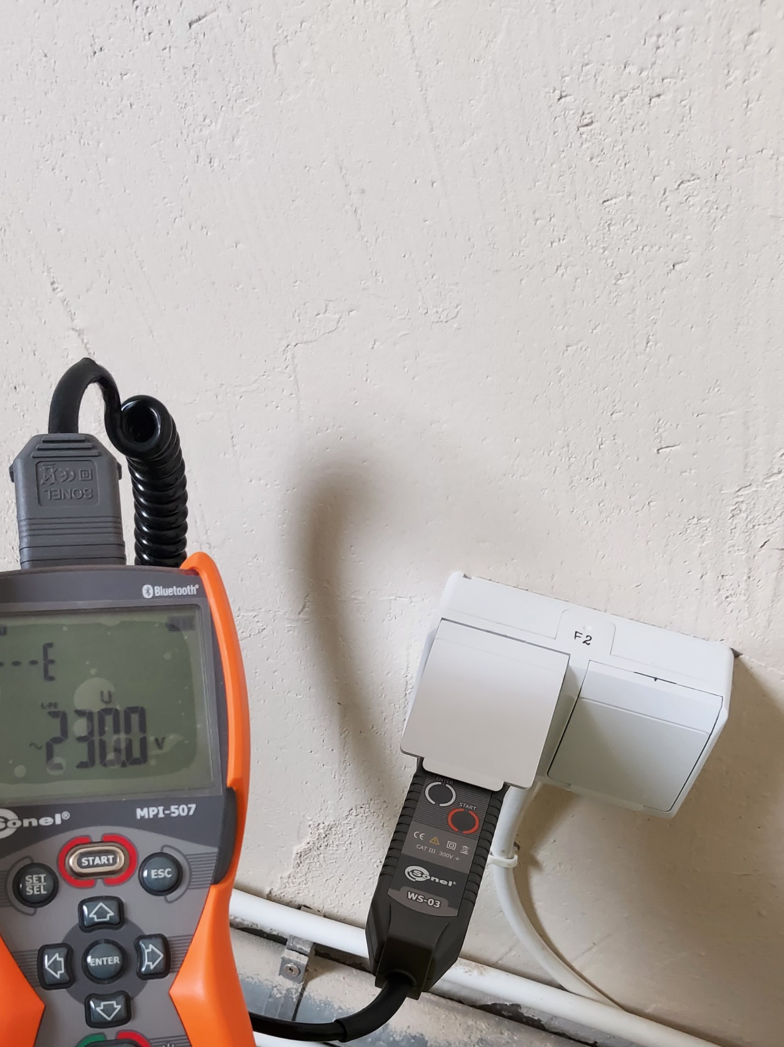 Miernik Sonel MPI-507 z pomarańczową obudową wyświetlający napięcie 230.0 V, podłączony do gniazdka elektrycznego za pomocą czarnego adaptera Sonel WS-03, na tle ściany o nierównej fakturze.
