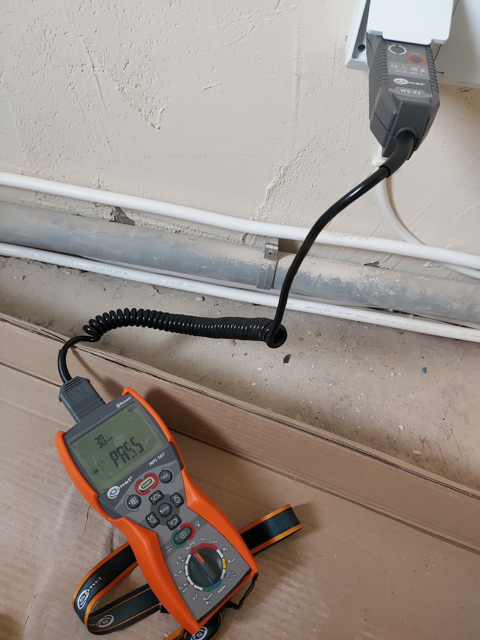 Miernik elektryczny Sonel MPI-507 z napisem 'PASS' na ekranie, podłączony do gniazdka elektrycznego za pomocą czarnego kabla spiralnego, leżący na kartonowej podłodze. W tle widoczne rury...