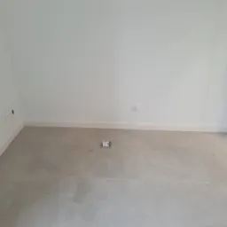 Pusty, jasny pokój z szarą podłogą wyłożoną płytkami, białe ściany z listwami przypodłogowymi i gniazdami elektrycznymi w ścianie oraz gniazdem podłogowym z metalową ramką.