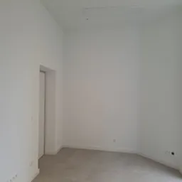 Puste, nowo wykończone pomieszczenie z szarą podłogą, białymi ścianami, listwami przypodłogowymi i gniazdkami elektrycznymi. Widoczny fragment okna po prawej stronie i wejście do drugiego...