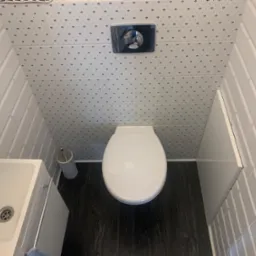 Nowoczesna toaleta z podwieszaną miską WC, umywalką narożną i ciemną podłogą imitującą drewno; ściany wyłożone białymi, prostokątnymi płytkami oraz płytkami z drobnym wzorem.