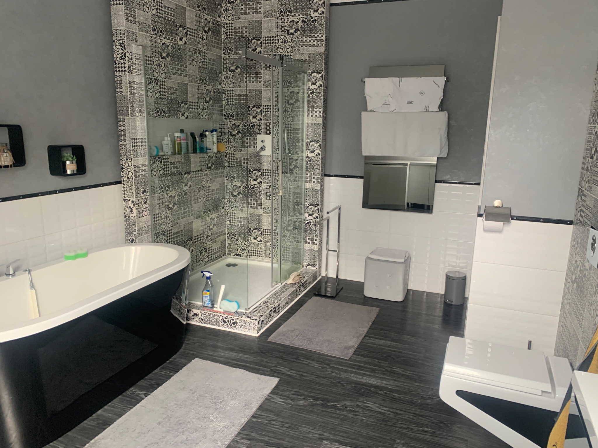 Nowoczesna łazienka z czarno-białymi wzorzystymi płytkami, wolnostojącą wanną na czarnych nóżkach, kabiną prysznicową ze szklanymi drzwiami, ciemną podłogą imitującą drewno i białymi elementami...