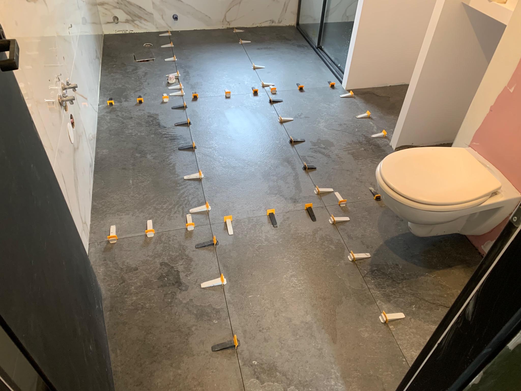 Układanie ciemnych płytek podłogowych w łazience w Trzebini, z użyciem systemu poziomowania płytek (klipsy i kliny) na świeżo nałożonym kleju, widoczna miska ustępowa podwieszana.