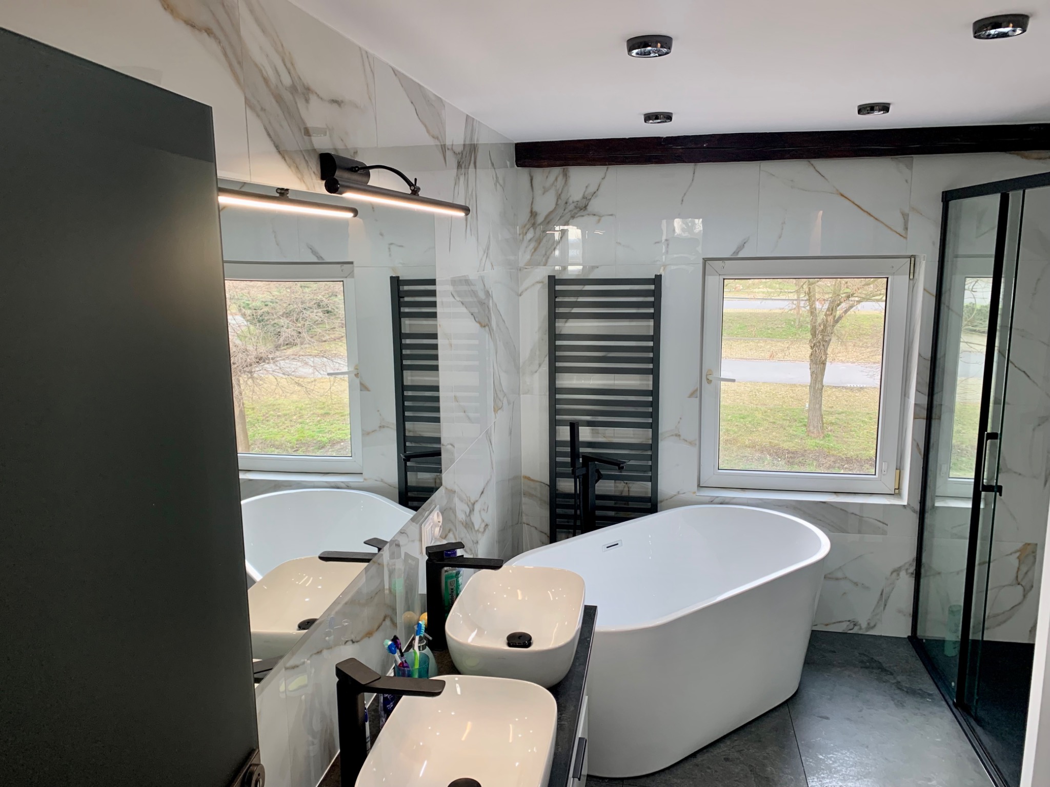 Nowoczesna łazienka z marmurowymi płytkami, wolnostojącą wanną, dwiema umywalkami nablatowymi i czarnymi akcentami, widok na zewnątrz przez okno.