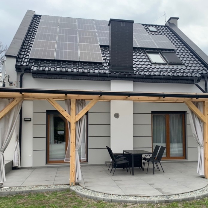 Elewacja domu z panelami słonecznymi na dachu, taras z drewnianą pergolą i zasłonami, szara kostka brukowa i meble ogrodowe.