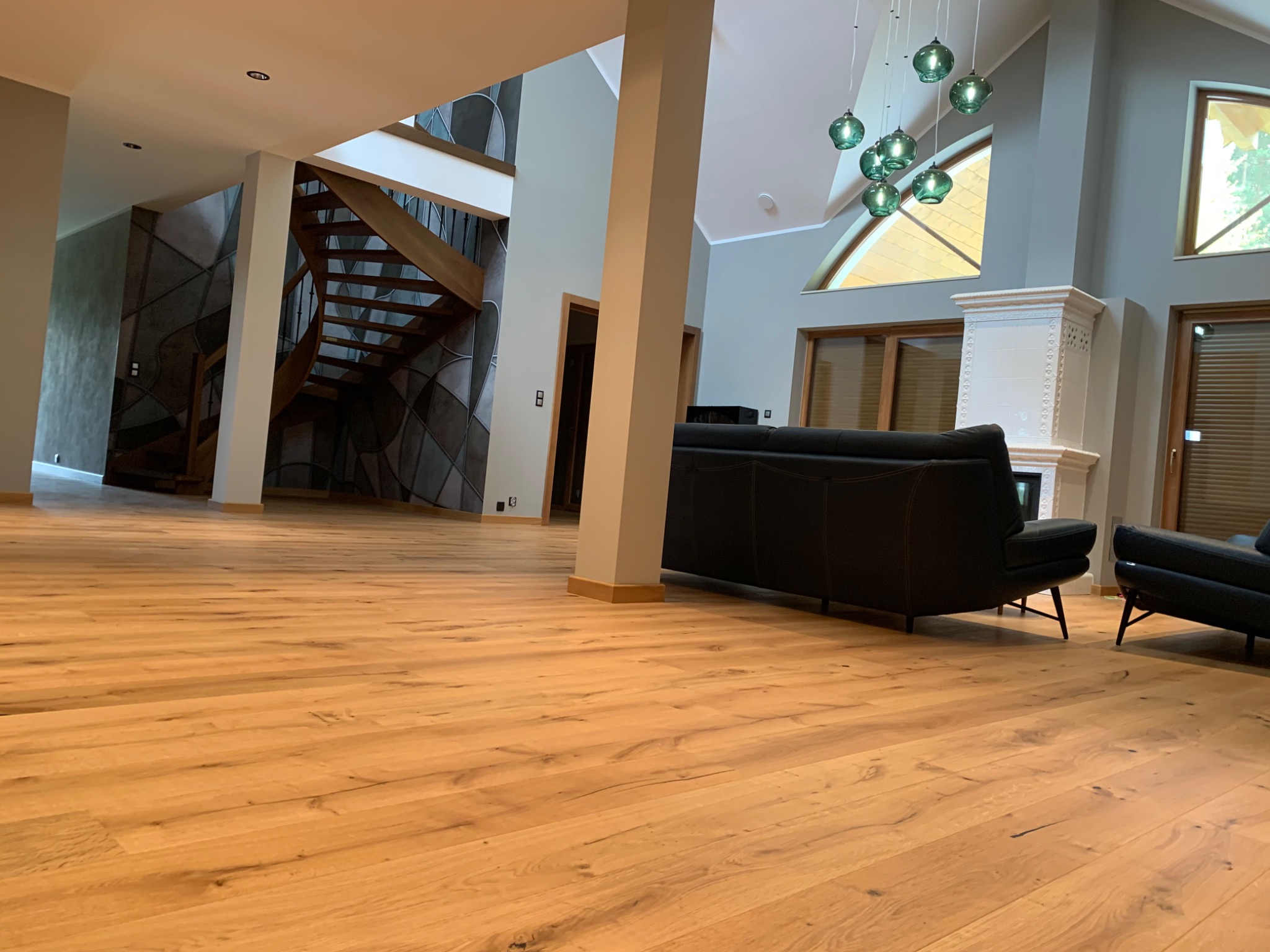 Rozległy salon z nową, jasną podłogą z desek, nowoczesnymi meblami i schodami w tle, oświetlony wiszącymi lampami i światłem dziennym.